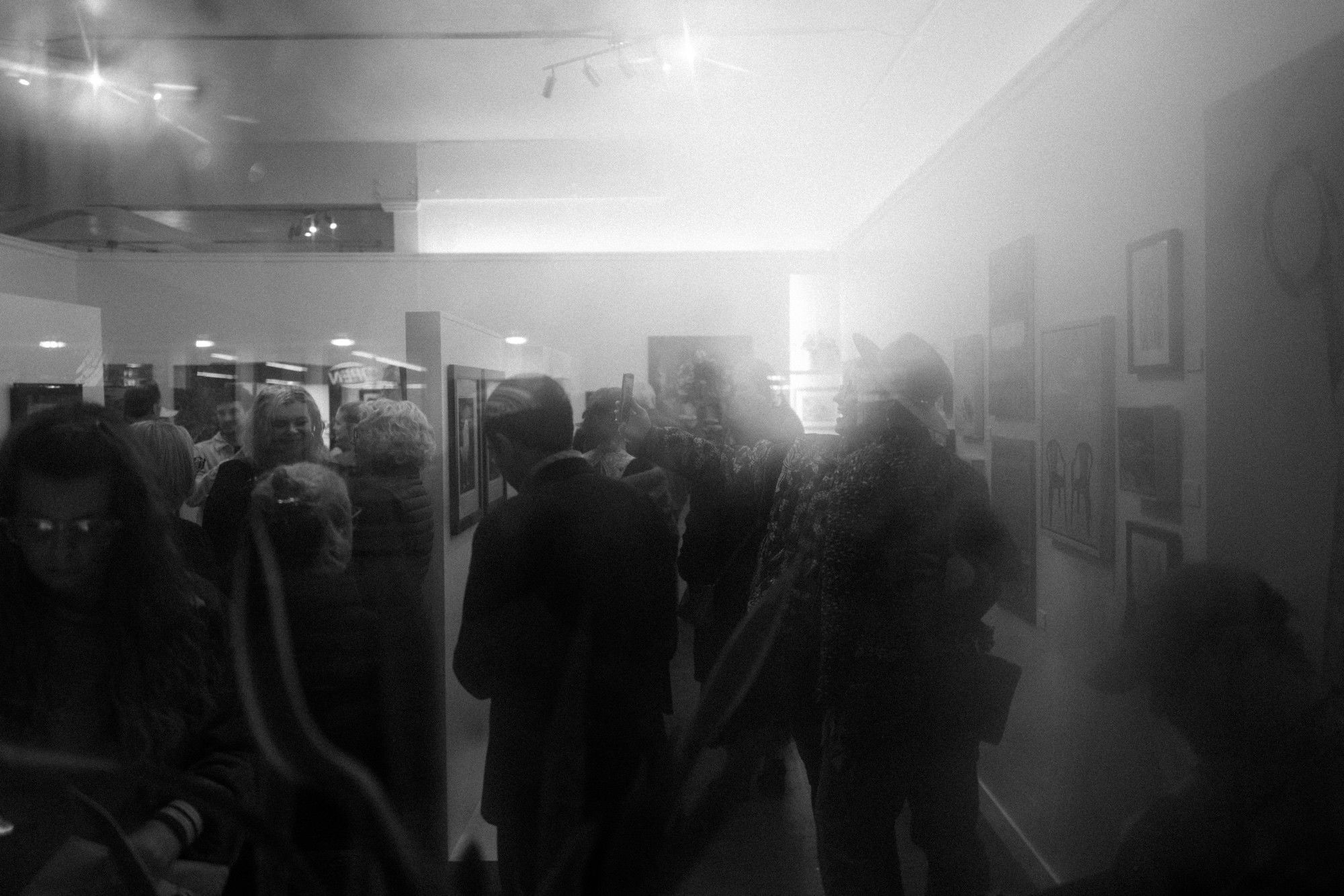 Visitors mingling at a gallery event, some taking selfies, with lens flare from overhead lights.