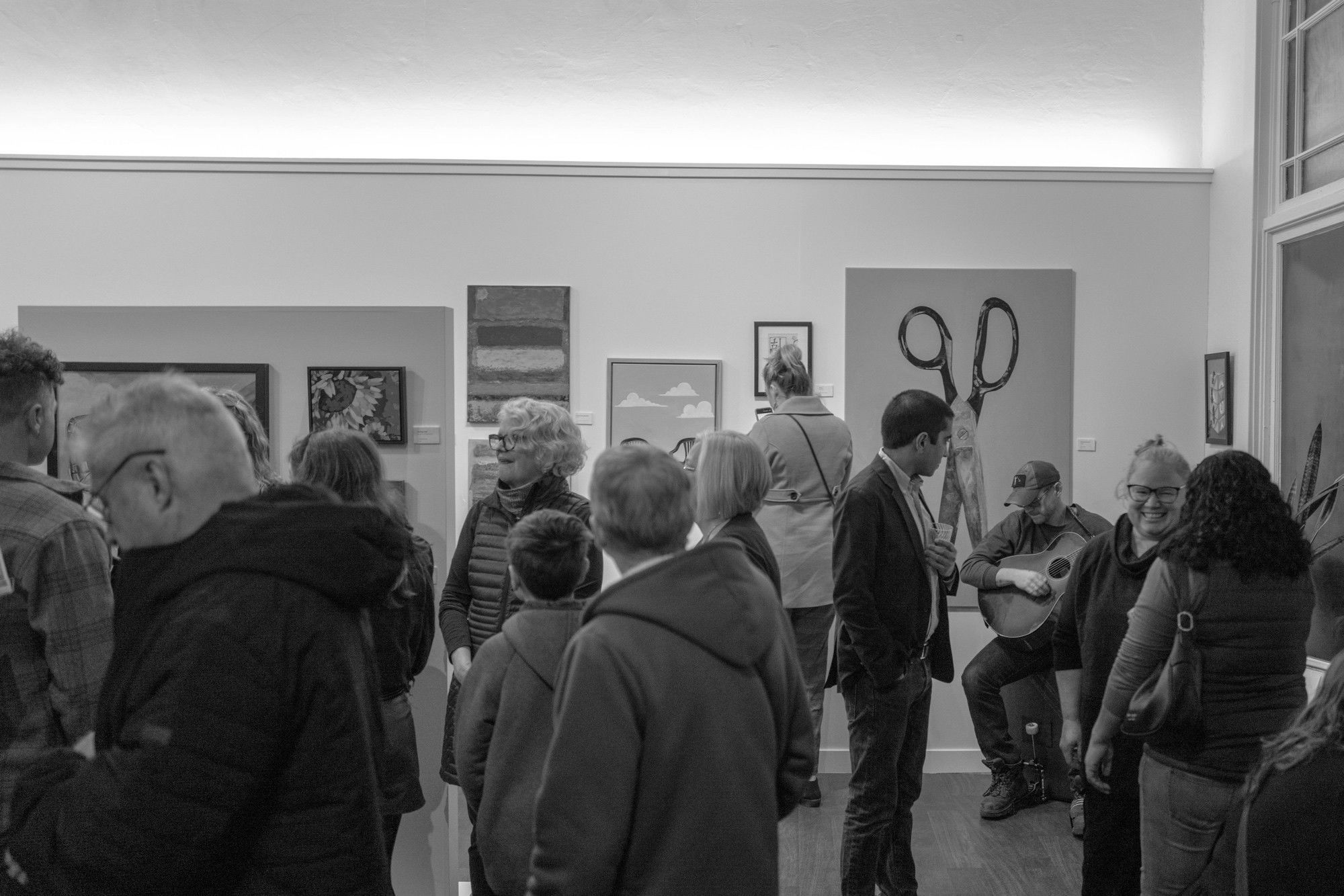 Visitors mingle in gallery with paintings displayed on walls, while a seated musician plays acoustic guitar.
