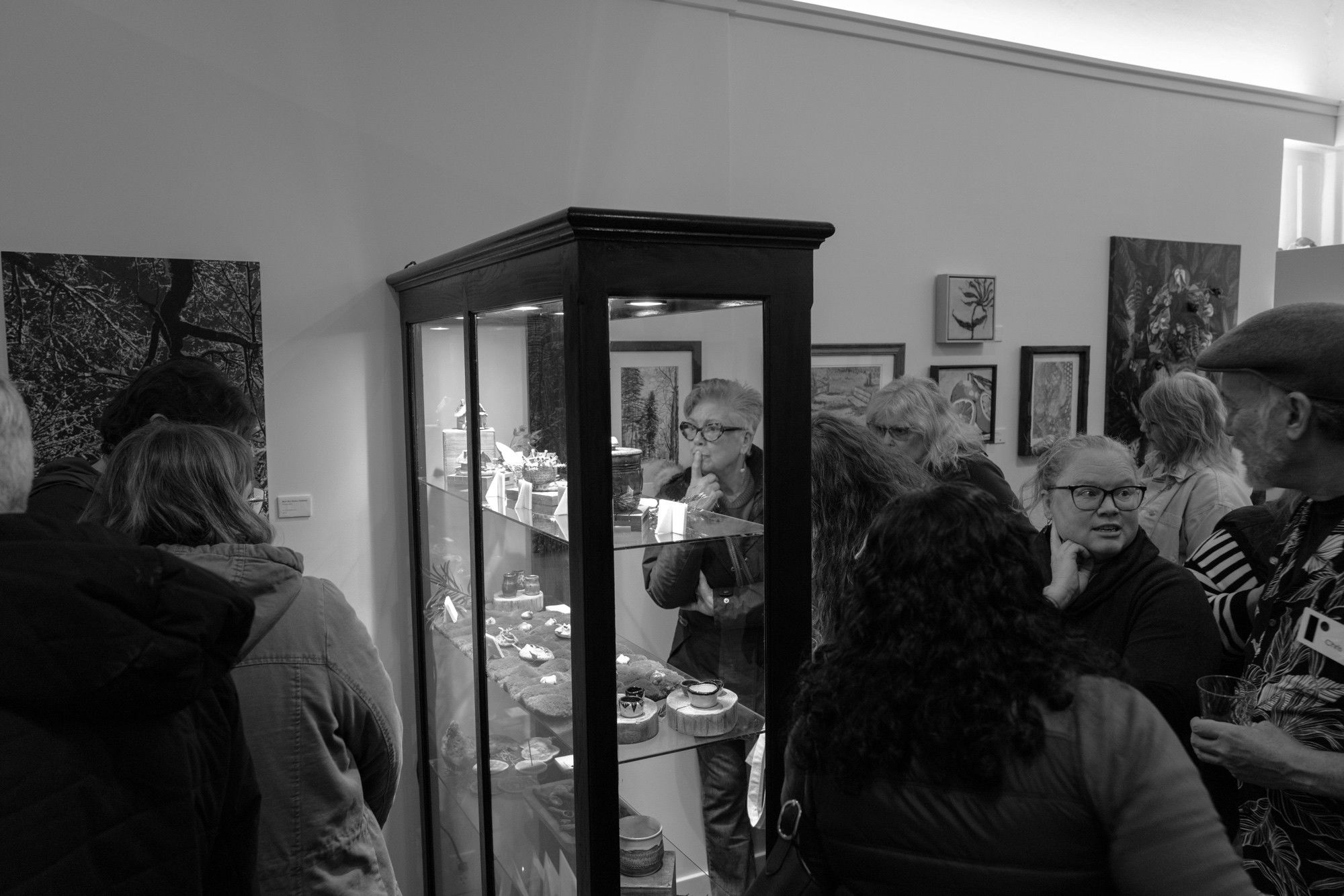 Gallery visitors converse around a tall glass display case showcasing ceramics and small artworks, with framed pieces on walls behind.